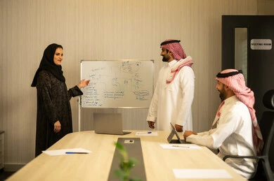 Using a tablet for note-taking, a Saudi Gulf Arab team consisting of two men wearing traditional thob and ghutrah and a hijab-wearing woman in an abaya explaining to her colleagues using a whiteboard, teamwork and collaboration, discussing a training and development project, clarifying the processes and procedures followed in the company.
