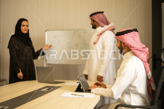 العمل الجماعي و التعاون، مناقشة مشروع تدريب و تطوير، توضيح العمليات و الاجراءات المتبعة في الشركة، فريق عربي خليجي سعودي مكون من رجلين يرتديان الثوب التقليدي و الشماغ و سيدة محجبة ترتدي العباءة تشرح لزملائها باستعمال السبورة, استخدام التابلت في تدوين الملاحظات