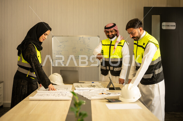 توضيح مخطط البناء، مفهوم التخطيط والتنظيم، متابعة سير المشروع، التعاون المهني داخل المكتب الهندسي، مهندس عربي خليجي سعودي يرتدي الشماغ و سترة السلامة يقف برفقة زملائه يقوم بشرح العمل
