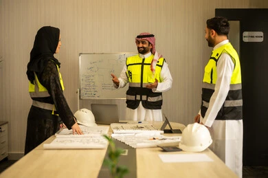 Study of the construction plan, the concept of planning and organization, monitoring the progress of the project, professional collaboration within the engineering office, a Saudi Gulf Arab engineer wearing a ghutrah and safety vest stands with his colleagues explaining the work.