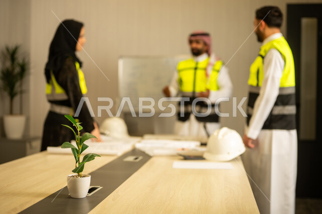 مراجعة مخطط البناء، مفهوم التخطيط والتنظيم، متابعة سير المشروع، التعاون المهني داخل المكتب الهندسي، مهندس عربي خليجي سعودي يرتدي الشماغ و سترة السلامة يقف برفقة زملائه يقوم بشرح العمل