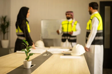 مراجعة مخطط البناء، مفهوم التخطيط والتنظيم، متابعة سير المشروع، التعاون المهني داخل المكتب الهندسي، مهندس عربي خليجي سعودي يرتدي الشماغ و سترة السلامة يقف برفقة زملائه يقوم بشرح العمل