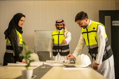 Monitoring the progress of the project, discussing the construction plan, professional collaboration within the engineering office, the concept of planning and organization, a Saudi Gulf Arab engineer wearing a ghutrah and safety vest stands with his colleagues explaining the work.