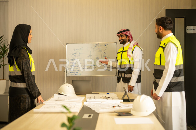 التعاون المهني داخل المكتب الهندسي، مناقشة مخطط البناء، متابعة سير المشروع، مفهوم التخطيط والتنظيم، مهندس عربي خليجي سعودي يرتدي الشماغ و سترة السلامة يقف برفقة زملائه يقوم بشرح العمل