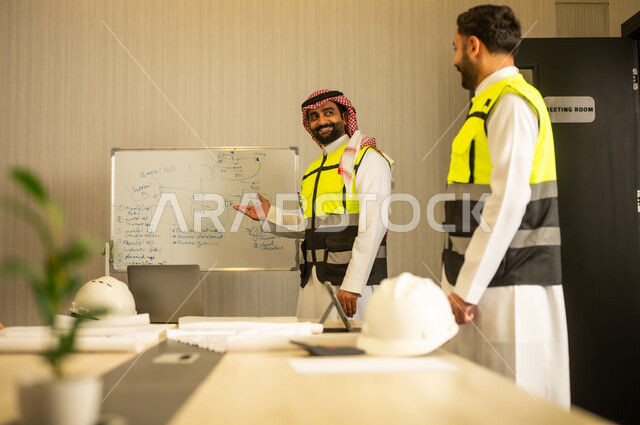 مفهوم التعاون المهني داخل بيئة العمل, اجتماع هندسي لمراجعة المخططات, مهندسيان عربيان خليجيان سعوديان يرتديان الزي التقليدي و سترة الحماية يناقشان سير العمل
