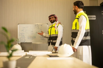 The concept of professional collaboration within the work environment, an engineering meeting to review plans, two Arab Gulf engineers from Saudi Arabia wearing traditional attire and safety vests discussing the workflow.