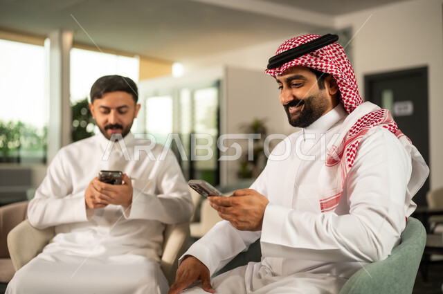 ايماءات الاندماج و التركيز، تصفح مواقع التواصل الاجتماعي عبر الانترنت، التواصل والدردشة مع الأصدقاء والأقارب عبر الجوال، استخدام جهاز تقني حديث ومتطور، رجلان عربيان خليجيان سعوديان يرتديان الزي التقليدي  يقومان باستخدام لهاتف المحمول