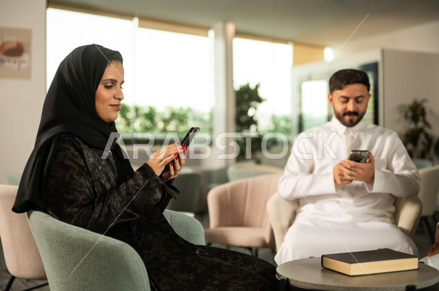 انجاز مهام العمل، استخدام جهاز تقني حديث، دمج التكنولوجيا في الحياة اليومية ، رجل عربي خليجي سعودي يرتدي الثوب الابيضيجلس برفقة زميلته يحمل بيده هاتف محمول، تصفح مواقع و وسائل التواصل الاجتماعي