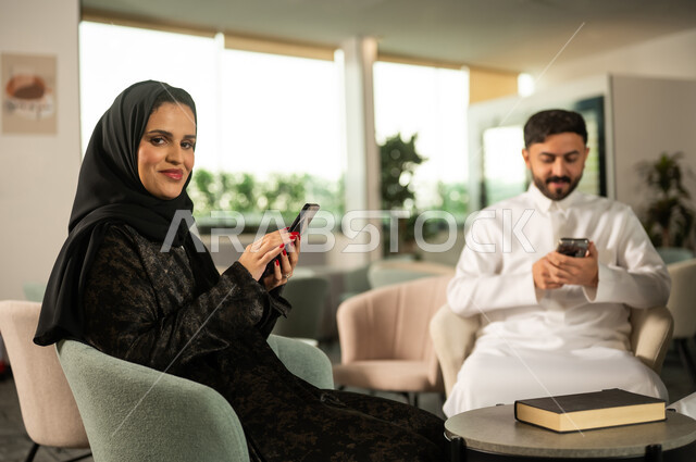 استخدام جهاز تقني حديث ، انجاز مهام العمل، دمج التكنولوجيا في الحياة اليومية ، رجل عربي خليجي سعودي يرتدي الثوب الابيضيجلس برفقة زميلته يحمل بيده هاتف محمول، تصفح مواقع و وسائل التواصل الاجتماعي