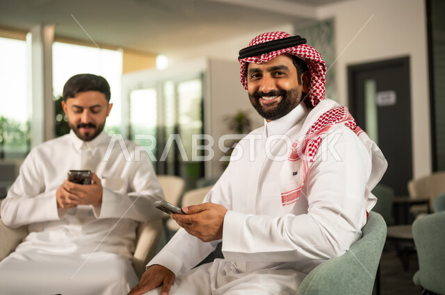 تصفح مواقع التواصل الاجتماعي عبر الانترنت، التواصل والدردشة مع الأصدقاء والأقارب عبر الجوال، استخدام جهاز تقني حديث ومتطور، رجلان عربيان خليجيان سعوديان يرتديان الزي التقليدي  يقومان باستخدام لهاتف المحمول