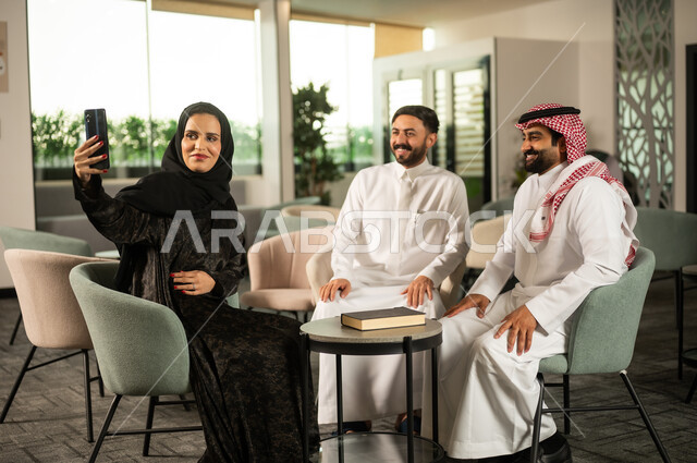 تطوير الخطط وتحقيق أهداف الشركة، التقاط صور السيلفي التذكارية عبر الجوال، الحوارات و النقاشات حول المشاريع، التعاون و المشاركة في إنجاز الأعمال، فريق عمل عربي خليجي سعودي مكون من رجلين يرتديان الزي التقليدي  و سيدة محجبة ترتدي عباءة  ينظروا تمسك بيديها الهاتف المحمول