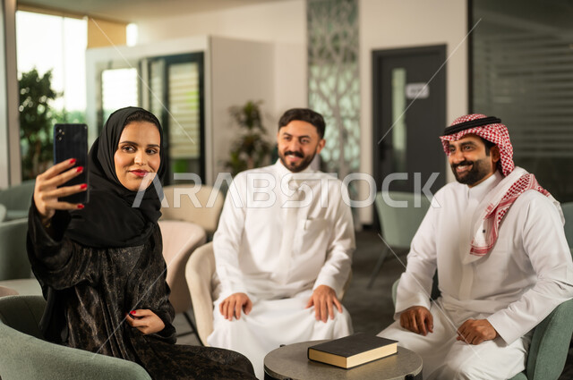 التقاط صور السيلفي التذكارية عبر الجوال، الحوارات و النقاشات حول المشاريع، تطوير الخطط وتحقيق أهداف الشركة، التعاون و المشاركة في إنجاز الأعمال، فريق عمل عربي خليجي سعودي مكون من رجلين يرتديان الزي التقليدي  و سيدة محجبة ترتدي عباءة  ينظروا تمسك بيديها الهاتف المحمول