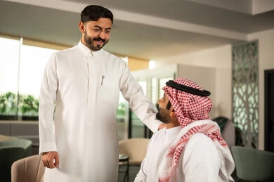 Gaining experience and expanding the professional network, a Saudi Gulf Arab man wearing a white thobe is talking to a colleague, collaboration and teamwork, directing efforts to achieve the company's goals.