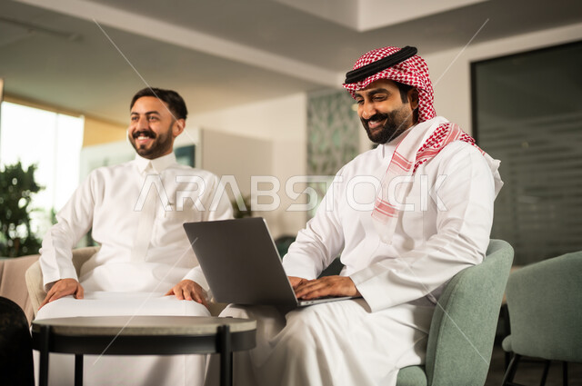 ايماءات الاندماج و التركيز، متابعة مهمة عمل عبر حاسب محمول، مفهوم التعاون المهني، موظف عربي خليجي سعودي يرتدي الثوب و الشماغ يجلس أمام حاسب محمول برفقة زميلة، أجواء عمل إيجابية
