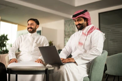 Gestures of integration and focus, following a work task via a laptop, the concept of professional collaboration, a Saudi Gulf Arab employee wearing a thobe and shemagh sitting in front of a laptop with a colleague, positive work atmosphere.
