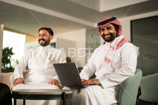النظر للكاميرا بتعابير السعادة و السرور، متابعة مهمة عمل عبر حاسب محمول، مفهوم التعاون المهني، موظف عربي خليجي سعودي يرتدي الثوب و الشماغ يجلس أمام حاسب محمول برفقة زميلة، أجواء عمل إيجابية