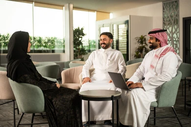 Gestures of happiness and joy, exchanging ideas and discussing daily tasks, teamwork and professional communication, two Arab Gulf Saudi employees wearing traditional attire discussing work content alongside their colleague via a laptop.