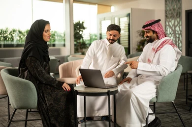 تبادل الأفكار ومناقشة المهام اليومية، العمل الجماعي والتواصل المهني, موظفان عربيان خليجيان سعوديان يرتديان الزي التقليدي يناقشان محتوى العمل برفقة زميلتهم عبر الحاسوب المحمول، الاشارة الى شاشة الابتوب