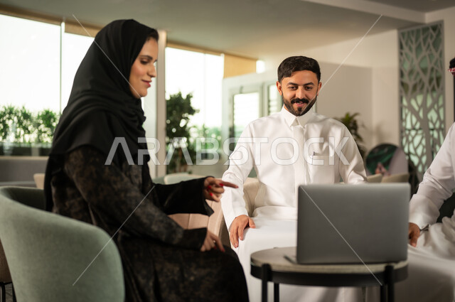 Collaboration within the work environment, communication and coordination of tasks among colleagues, an Arab Gulf Saudi female employee in an abaya and hijab while interacting with the team, using a laptop during the meeting, pointing to the laptop screen.