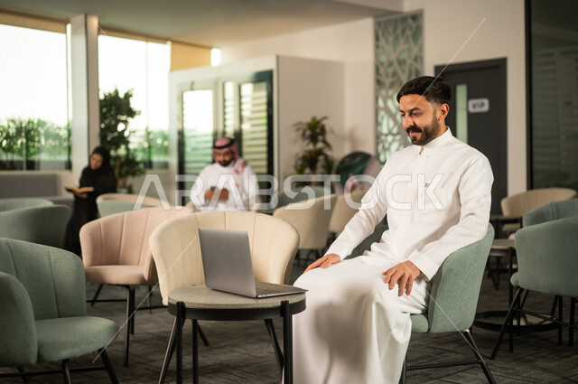 اجراء مكالمة فيديو ، إنجاز العمل اونلاين من خلال الحاسب المحمول، رجل عربي خليجي سعودي يرتدي الثوب الابيض يجلس على كرسي امام الابتوب، تطور التكنولوجيا وإستخدام الأجهزة التقنية الحديثة