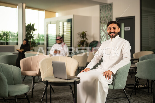 النظر الى الكاميرا بتعابير السرور، إنجاز العمل اونلاين من خلال الحاسب المحمول، رجل عربي خليجي سعودي يرتدي الثوب الابيض يجلس على كرسي امام الابتوب، تطور التكنولوجيا وإستخدام الأجهزة التقنية الحديثة