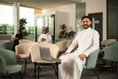 Looking at the camera with expressions of joy, accomplishing work online through a laptop, an Arab Gulf Saudi man wearing a white thobe sitting on a chair in front of the laptop, the development of technology and the use of modern technical devices.