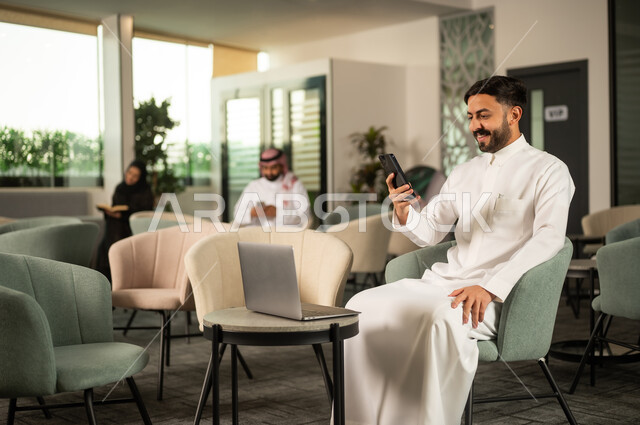 انجاز المهام عن بعد، تصفح تطبيقات الهاتف المحمول, استخدام جهاز تقني حديث ، دمج التكنولوجيا في الحياة اليومية ، رجل عربي خليجي  سعودي يرتدي الثوب الابيض يجلس على كرسي امام الحاسب المحمول يحمل بيده الجوال وينظر بايماءات السعادة ، تصفح مواقع و وسائل التواصل الاجتماعي