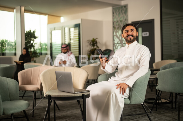 تصفح تطبيقات الهاتف المحمول, استخدام جهاز تقني حديث ، انجاز المهام عن بعد، دمج التكنولوجيا في الحياة اليومية ، رجل عربي خليجي  سعودي يرتدي الثوب الابيض يجلس على كرسي امام الحاسب المحمول يحمل بيده الجوال وينظر الى شئ ما بايماءات السعادة ، تصفح مواقع و وسائل التواصل الاجتماعي
