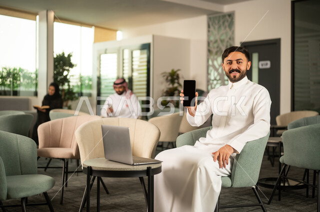 استخدام الأجهزة والتقنيات الحديثة، مهن و أعمال إدارية مكتبية، رجل عربي خليجي سعودي يرتدي الثوب الابيض يجلس على كرسي امام الحاسب المحمول يمسك بيده الهاتف المحمول يقوم بعرض شاشة سوداء فارغة, ايماءات السعادة و السرور