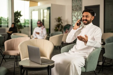 إجراء مكالمة هاتفية صوتية عبر الجوال، الاستمتاع بتبادل الأحاديث و الأخبار،رجل عربي خليجي سعودي يرتدي الثوب الابيض يجلس امام الحاسب المحمول يتحدث عبر الهاتف المحمول، إيماءات السعادة بالتواصل مع الأصدقاء والأقارب