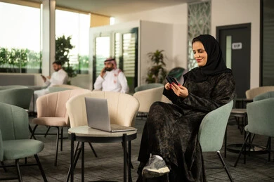 قضاء وقت ممتع عبر الهاتف المحمول، تصفح موقع التواصل الاجتماعي، امراة عربية خليجية سعودية محجبة ترتدي العباءة تجلس على كرسي امام الحاسب المحمول تحمل بيدها الجوال تنظر بإيماءات السرور