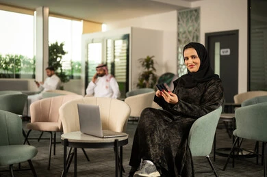 Browsing social media, a Saudi Arabian Gulf woman in a hijab wearing an abaya sits on a chair in front of a laptop, holding a mobile phone in her hand, looking with gestures of joy, enjoying her time on the mobile phone.