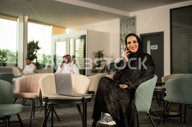 Sharing news and conversations, communicating with family and friends, using modern technological devices in daily life, a Saudi Arabian Gulf woman wearing a hijab and an abaya sitting in front of a laptop making a phone call via a mobile phone, gestures of happiness and joy.
