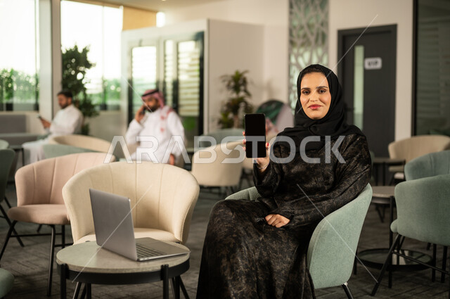 عرض شاشة سوداء فارغة عبر الهاتف المحمول، استخدام جهاز تقني حديث ومتطور، امرأة عربية خليجية سعودية محجبة تجلس امام الحاسب المحمول تحمل بيدها الجوال، النظر الى الكاميرا بايماءات السعادة والسرور