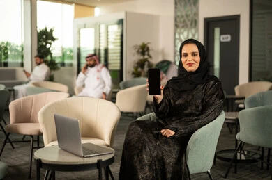 عرض شاشة سوداء فارغة عبر الهاتف المحمول، استخدام جهاز تقني حديث ومتطور، امرأة عربية خليجية سعودية محجبة تجلس امام الحاسب المحمول تحمل بيدها الجوال، النظر الى الكاميرا بايماءات السعادة والسرور