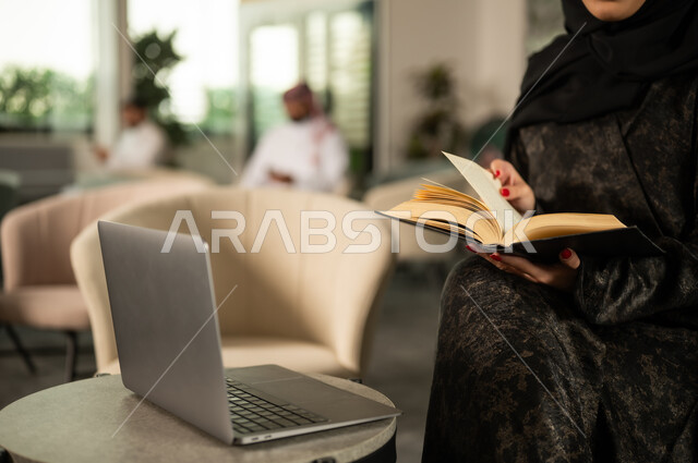 The concept of personal development and knowledge, combining reading and technology in daily work, a close-up image of a Saudi Gulf Arab woman sitting in front of a laptop and reading a book, the culture of self-learning.