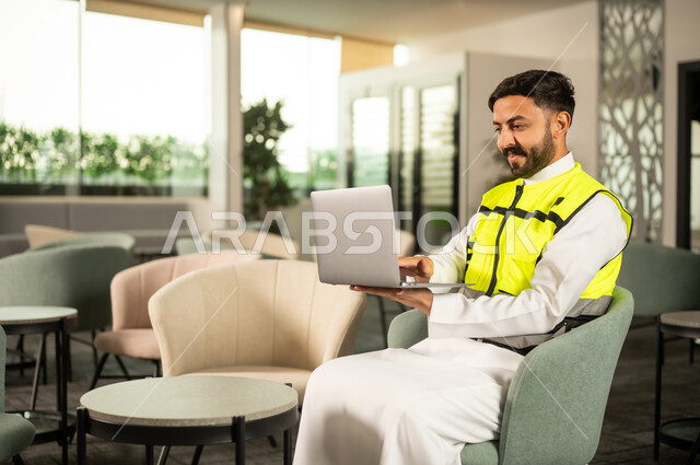 Organizing and monitoring work using a laptop, using technology in engineering supervision, the concept of modern architecture, an Arab Gulf Saudi engineer wearing traditional attire and a safety vest working on a laptop.
