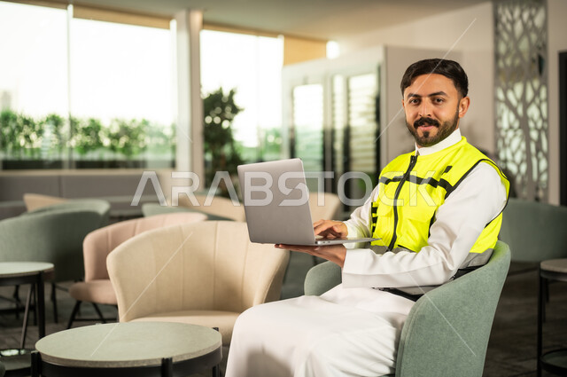 متابعة المهام باستخدام الحاسب المحمول، استخدام التكنولوجيا في الإشراف الهندسي، مفهوم الهندسة المعمارية الحديثة، متابعة تنفيذ المشروع الهندسي داخل المكتب, مهندس عربي خليجي سعودي يرتدي الثوب الابيض وسترة السلامة يعمل على الابتوب