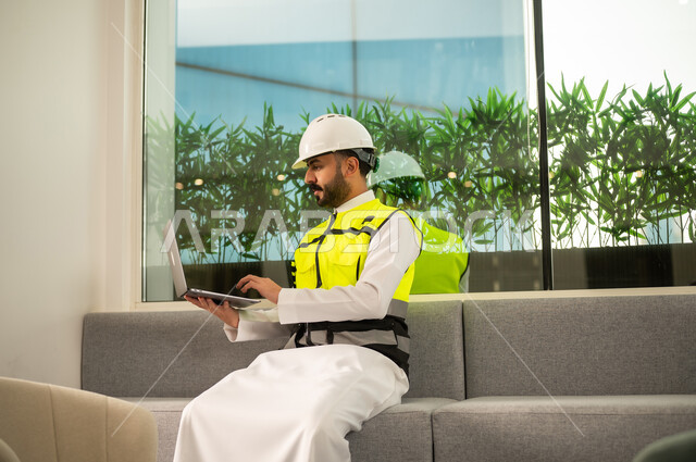 استخدام التكنولوجيا في الإشراف الهندسي، مفهوم الهندسة المعمارية الحديثة، متابعة تنفيذ المشروع الهندسي داخل المكتب, مهندس عربي خليجي سعودي يرتدي خوذة وسترة السلامة يعمل على الحاسب المحمول