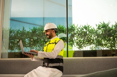 مفهوم الهندسة المعمارية الحديثة، متابعة تنفيذ المشروع الهندسي داخل المكتب, استخدام التكنولوجيا في الإشراف الهندسي, مهندس عربي خليجي سعودي يرتدي خوذة وسترة السلامة يعمل على الحاسب المحمول