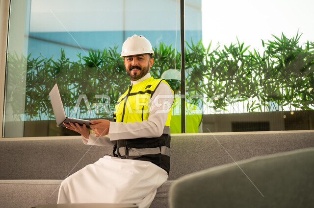 متابعة تنفيذ المشروع الهندسي داخل المكتب، النظر الى الكامير بتعابير السرور, مفهوم الهندسة المعمارية الحديثة, استخدام التكنولوجيا في الإشراف الهندسي, مهندس عربي خليجي سعودي يرتدي خوذة وسترة السلامة يحمل الحاسب المحمول