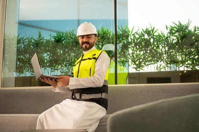 متابعة تنفيذ المشروع الهندسي داخل المكتب، النظر الى الكامير بتعابير السرور, مفهوم الهندسة المعمارية الحديثة, استخدام التكنولوجيا في الإشراف الهندسي, مهندس عربي خليجي سعودي يرتدي خوذة وسترة السلامة يحمل الحاسب المحمول