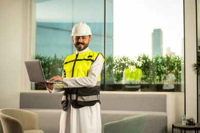 النظر الى الكامير بتعابير السرور، متابعة تنفيذ المشروع الهندسي داخل المكتب, مفهوم الهندسة المعمارية الحديثة, استخدام التكنولوجيا في الإشراف الهندسي, مهندس عربي خليجي سعودي يرتدي خوذة وسترة السلامة يحمل الحاسب المحمول
