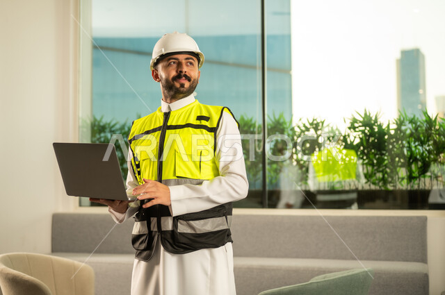 Monitoring the implementation of the engineering project inside the office, the concept of modern architecture, the use of technology in engineering supervision, an Arab Gulf Saudi engineer wearing a helmet and safety vest holding a laptop looking at something.