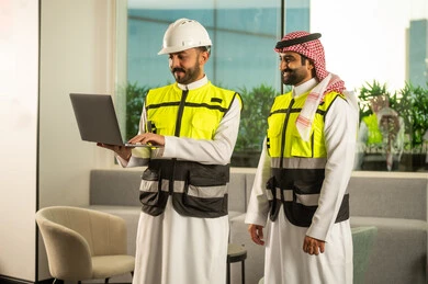 The concept of supervision and follow-up, management of engineering projects, professional coordination within the work environment, a Saudi Gulf Arab engineer wearing a helmet and safety vest reviewing data on a laptop and discussing work steps with a colleague.