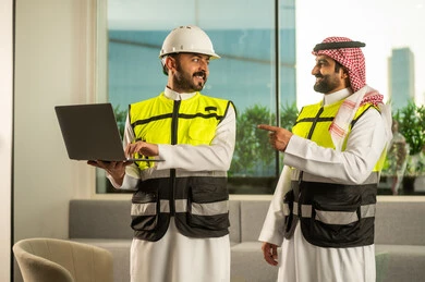 إدارة المشاريع الهندسية, مفهوم التخطيط والتنفيذ, التنسيق المهني داخل بيئة العمل, مهندس عربي خليجي سعودي يرتدي خوذة و سترة الحماية يراجع البيانات على الحاسب المحمول و يناقش خطوات العمل برفقة زميلة