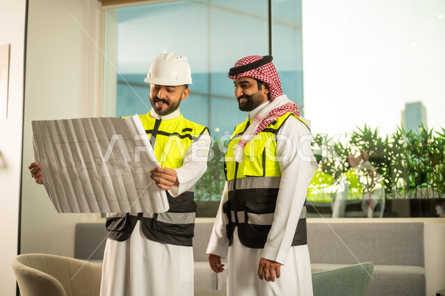التخطيط ووضع اساسيات المخططات المعمارية، متابعة تنفيذ الخطط الهندسية، الشركات المختصة بورشات البناء والمقاولات، مهندسان عربيان خليجيان سعوديان يرتديان سترة الحماية يعملان على مناقشة مخطط الإنشاء وتحديد القياسات بدقة