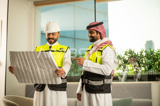 Referring to something, following up on the implementation of engineering plans, companies specialized in construction workshops and contracting, planning and establishing the fundamentals of architectural designs, two Arab Gulf engineers from Saudi Arabia wearing safety vests are discussing the construction plan and accurately determining the measurements.