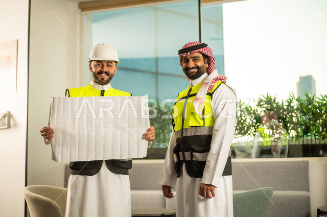 النظر الى الكاميرا بتعابير السرور، متابعة تنفيذ الخطط الهندسية، الشركات المختصة بورشات البناء والمقاولات، التخطيط ووضع اساسيات المخططات المعمارية، مهندسان عربيان خليجيان سعوديان يرتديان سترة الحماية يعملان على مناقشة مخطط الإنشاء وتحديد القياسات بدقة