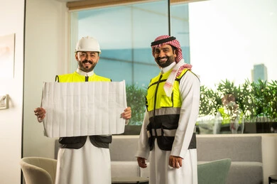 Looking at the camera with expressions of joy, following the implementation of engineering plans, companies specialized in construction workshops and contracting, planning and establishing the fundamentals of architectural designs, two Arab Gulf engineers from Saudi Arabia wearing safety vests are discussing the construction plan and accurately determining the measurements.